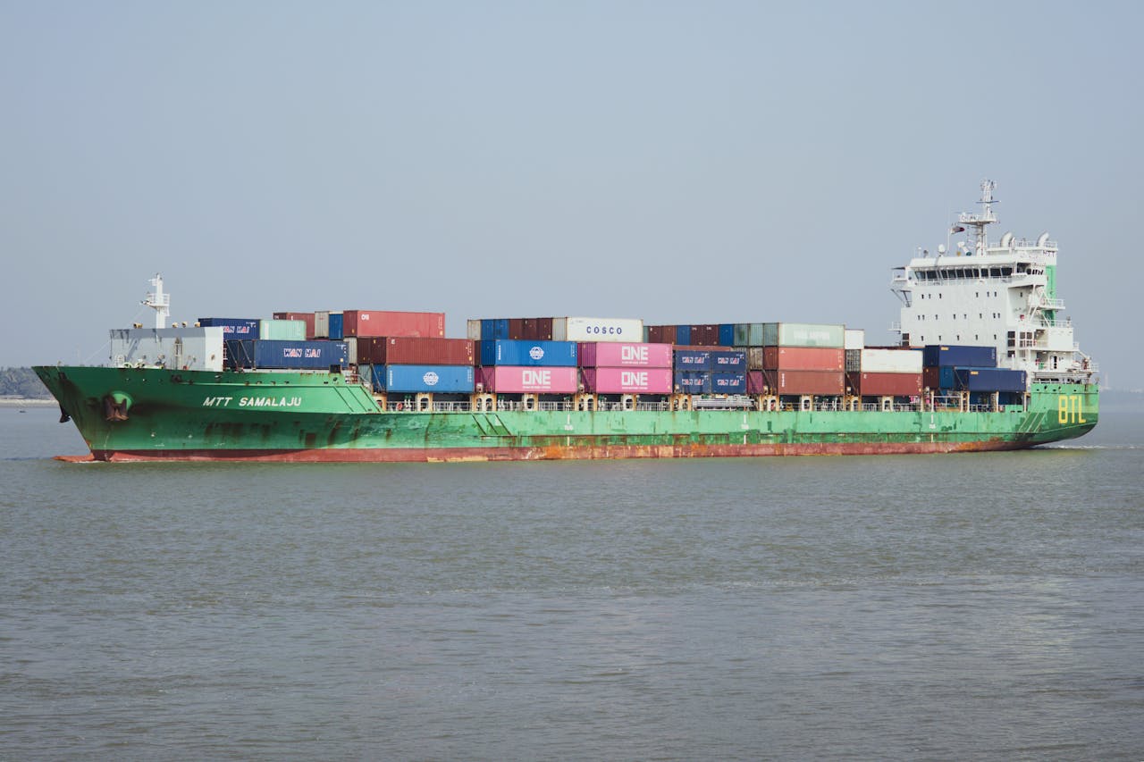 A cargo ship carrying multiple containers navigates through calm waters under a clear blue sky.